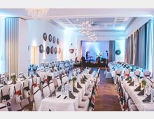 Großer Veranstaltungssaal mit weiß gedeckten Tischen und Stühlen, die in Reihen angeordnet sind. Im Hintergrund eine Bühne mit Musikern, seitlich mit Vorhängen und an den Wänden runde Bilderrahmen im klassischen Stil.