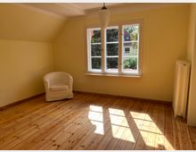 Ein leerer Raum mit gelben Wänden, Holzdielenboden und einem großen Fenster mit weißem Rahmen, das Tageslicht hereinscheinen lässt. Ein weißer Sessel steht in der linken Ecke, und rechts neben dem Fenster befindet sich ein Heizkörper.