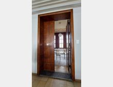 Holzrahmentür mit teilweise geöffnetem Flügel, die zu einem hellen Raum führt. Im Inneren sind große Fenster, einfache weiße Tische und Stühle sowie eine Hängelampe sichtbar.