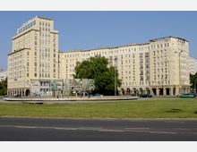 Ein großes, mehrstöckiges Wohn- und Bürogebäude im klassischen sozialistischen Baustil mit hellen Fassaden und regelmäßigen Fensterreihen, umgeben von einer Grünanlage mit einem Brunnen und modernen Skulpturen im Vordergrund. Der Himmel ist wolkenlos, und einige Autos sind auf der Straße zu sehen.