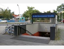 Eingang zur U-Bahn-Station "Konstanzer Straße" mit blauer Stationsbeschilderung, einer Treppe, die nach unten führt, und mehreren abgestellten Fahrrädern links daneben. Im Hintergrund Bäume, parkende Autos und ein Fußweg.