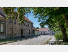 Straßenszene in einer Wohngegend mit Kopfsteinpflaster, links eine Reihe älterer Häuser mit verputzten Fassaden und geschlossenen Fensterläden, rechts moderne Gebäude hinter Bäumen. Der Bereich ist von grünem Laub und Schatten überspannt.