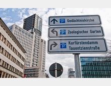 Straßenschild in einer städtischen Umgebung mit Wegweisern zu „Gedächtniskirche“, „Zoologischer Garten“ und „Kurfürstendamm/Tauentzienstraße“, vor modernen Bürogebäuden und einem Hochhaus. Der Himmel ist teilweise bewölkt.