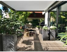 Moderne Terrasse mit Holzboden, einer Pergola mit weißem Lamellendach und großen Glas-Schiebetüren. Die Terrasse ist mit Pflanzen in dunklen Pflanzgefäßen, Sitzmöbeln und einem Esstisch ausgestattet, im Hintergrund ist ein Grill und ein Garten mit Bäumen sichtbar.