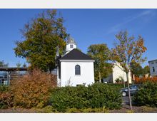 Weiße, kleine Kapelle mit geschwungenem Fenster und dunkelgrauem Dach, im Vordergrund von Büschen und Bäumen umgeben. Im Hintergrund sind weitere Gebäude und ein klarer blauer Himmel sichtbar.