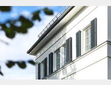 Weiße Fassade eines Gebäudes mit mehreren rechteckigen Fenstern, die von dunkelgrauen Fensterläden flankiert werden. Im oberen Bereich befindet sich ein Dachgesims mit einer schmalen Metallbrüstung, Teile von Zweigen ragen in den Bildvordergrund.