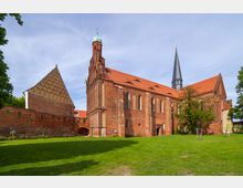 Historische Backsteinkirche mit roten Ziegeldächern, großen gotischen Fenstern und einem schlanken Turm mit Spitzhelm, umgeben von einer alten Ziegelmauer und einer grünen Rasenfläche im Vordergrund. Links ist ein Giebelgebäude mit auffälligem Muster sichtbar.