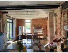 Offener Wohnraum mit industriellem Design; freiliegende Ziegelwände, schwarze Stahlträger an der Decke und ein polierter Betonboden. Im Raum befinden sich ein Esstisch mit Stühlen, ein Kühlschrank, große Glastüren und Fenster, die Tageslicht hereinlassen, sowie sichtbare Rohrleitungen an den Wänden.