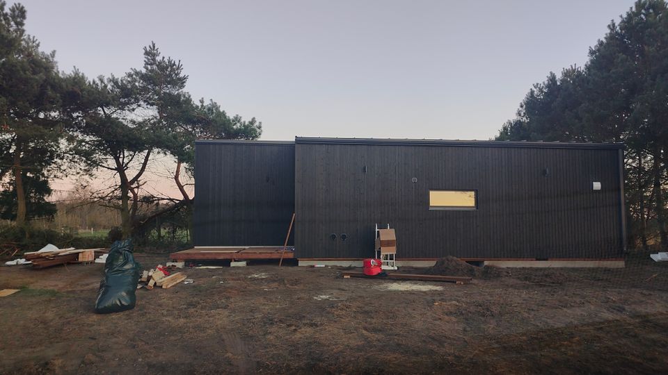 Ein langgestrecktes, modernes Gebäude mit schwarzer Holzfassade und flachem Dach steht auf einem sandigen Grundstück. Davor liegen Baumaterialien und Erdhaufen, umgeben von Kiefern und niedriger Vegetation.