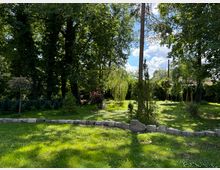 Grüne Parklandschaft mit verschiedenen Bäumen, including einer Weide und Kiefern, sowie einer locker gesetzten Steinreihe im Vordergrund. Im Hintergrund sind Büsche und dichte Vegetation unter einem teils bewölkten Himmel zu sehen.