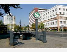 Straßenszene mit Eingang zur S-Bahn-Station "Anhalter Bahnhof", markiert durch ein grünes S-Bahn-Schild. Im Hintergrund befinden sich mehrstöckige Gebäude, darunter ein modernes Hochhaus und ein weißer Gebäudekomplex mit Geschäften und Cafés im Erdgeschoss.