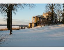 Schneebedeckte Landschaft mit einem historischen Gebäude, das von Bäumen umgeben ist. Das Gebäude hat eine Säulenveranda und liegt auf einer leichten Anhöhe, im Hintergrund ist ein See mit Schilf zu sehen.