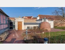 Ein kleiner Innenhof mit rot gepflastertem Boden, umgeben von historischen Gebäuden. Im Vordergrund befindet sich eine runde Backsteinkuppel, daneben eine moderne Tür eingebettet in einen weißen Gebäudeteil mit Backsteinwand; im Hintergrund sind Dächer und Fachwerkdetails sichtbar.