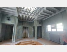 Innenraum mit hellgrünen, gefliesten Wänden und symmetrischen Säulen, die eine Nische mit einer Dusche oder Wasseranschluss umrahmen. Die Decke ist mit geometrischen Stufenmustern gestaltet, und an der rechten Wand befinden sich Fenster sowie ein Heizkörper.