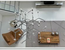 Minimalistisch eingerichtetes Wohnzimmer mit weißem Holzboden, einem modernen Kronleuchter aus Metall, einem cognacfarbenen Ledersessel, einem niedrigen Holztisch mit Dekorationen und einem strukturierten Teppich in Grautönen. Im Hintergrund ein helles Sideboard mit Fernseher.