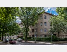 Mehrfamilienhaus aus der Nachkriegszeit mit hellbrauner Fassade und roten Ziegeldächern, umgeben von großen Bäumen. Im Vordergrund eine ruhige, von geparkten Autos gesäumte Straßenkreuzung mit Gehwegen und einem Straßenschild.