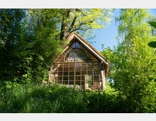 Ein kleines Holzhaus mit einer großen Glasfassade und einem bogenförmigen Fenster im Giebel, umgeben von dichtem grünen Laub und Bäumen unter einem klaren, blauen Himmel.