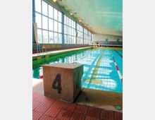 Innenansicht eines Schwimmbads mit einem Wettkampfbecken, das durch rot-weiße Trennleinen in Bahnen unterteilt ist. Links sind große Fensterfronten mit einfallendem Tageslicht zu sehen, und im Vordergrund steht ein Startblock mit der Zahl „4“.