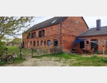 Backsteingebäude auf einem Hof mit Satteldach und mehreren Rundbogenfenstern, daneben ein kleiner Anbau mit überdachtem Sitzbereich und Holzmöbeln. Im Vordergrund ein Garten mit einem Zaun, Holzstapeln und einem Fahrrad, umgeben von Bäumen und Wiesen.