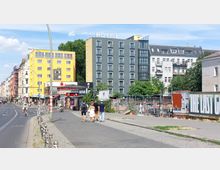 Straßenszene in einer Stadt mit mehrstöckigen Gebäuden, darunter ein modernes graues Hotel und gelbe Wohnhäuser. Im Vordergrund sind Gehwege, Geschäfte, ein Kiosk mit Imbissangeboten sowie einzelne Passanten und Radfahrer zu sehen; auf der rechten Seite eine Baustelle mit Graffiti und Plakaten.