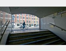 Blick aus einer mit weißen Fliesen verkleideten Unterführung mit Treppenstufen, die zu einer Straße führen, auf der ein Fahrradfahrer vorbeifährt; im Hintergrund ein großes Ziegelgebäude mit hohen Fenstern.