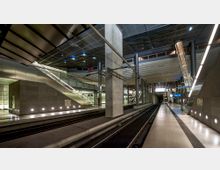 Moderne U-Bahn-Station mit breiten Bahnsteigen, mehrstöckigen Treppen und Rolltreppen sowie einer hohen Decke aus Metallplatten. Die Beleuchtung ist gleichmäßig, und die Wände bestehen teils aus Glas und Beton.
