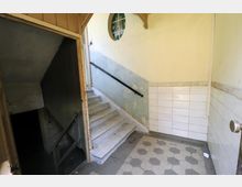 Treppe in einem alten Gebäude mit abgenutzten Wänden und Fliesen. Die Szenerie zeigt eine helle Wand mit einem runden Fenster, eine Treppe mit schwarzem Handlauf und eine dunkle Öffnung zu einem Kellerbereich.