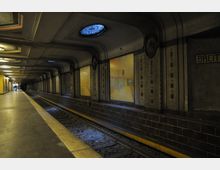 Unterirdischer U-Bahnhof mit abgerundeten Decken, dekorativen Wandverzierungen und einer durchgehenden Bahnsteigkante entlang der Gleise. Oberlichter lassen schwaches Licht herein, und ein Schild mit der Aufschrift "BREIT..." ist an der Wand angebracht.