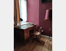 Holzschreibtisch mit einem Drehstuhl in einem Raum mit einer dunklen roten Wand; auf dem Tisch stehen eine Schreibtischlampe und ein Telefon. Daneben befindet sich eine Stehlampe mit passendem roten Lampenschirm, das Fenster ist mit weißen Vorhängen dekoriert, und der Boden besteht aus Holz.