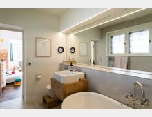 Modernes Badezimmer mit hellen Wänden, großem Spiegel und breitem Fenster. Im Vordergrund befinden sich eine freistehende Badewanne, ein Waschbecken auf einem Holzunterschrank und ein WC; im Hintergrund ist ein Blick in ein angrenzendes Zimmer mit Kinderaccessoires zu sehen.