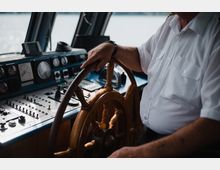 Ein Steuermann in Uniform hält das hölzerne Steuerrad eines Schiffs fest, umgeben von einem Bedienpult mit zahlreichen Schaltern, Instrumenten und Anzeigen, während im Hintergrund Wasser zu sehen ist.