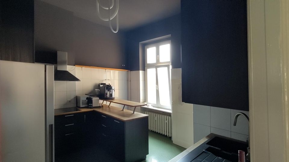 Schmale Küche mit dunklen Wänden, weißen Fliesen und schwarzen Unterschränken mit Holzarbeitsplatte. Ein großes Fenster über einem Heizkörper sorgt für Tageslicht; sichtbar sind zudem Kühlschrank, Dunstabzugshaube und Spüle.