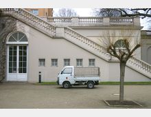 Gebäude mit beigefarbener Fassade, einer doppelläufigen Treppe mit Balustrade, großen Fenstern und vergitterten kleinen Fenstern; davor ein weißer Kleintransporter und ein Baum mit beschnittenen Ästen.