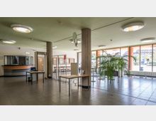 Heller Eingangsbereich eines öffentlichen Gebäudes mit Fliesenboden, großen Fenstern und mehreren Schreibtischen. Eine Sicherheitsschleuse, ein Empfangsschalter und eine große Zimmerpflanze sind sichtbar.