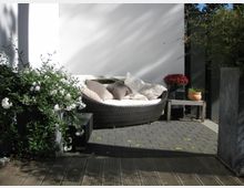 Moderner Außenbereich mit einer halbrunden Rattan-Liege, weißen Kissen und Pflanzen ringsum. Der Boden besteht aus Holz- und Steinflächen, und im Hintergrund steht eine weiße Mauer mit Blumentöpfen auf einem seitlichen Holztisch.