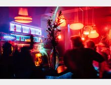 Ein Innenraum einer Bar mit bunter Beleuchtung in Rot- und Blautönen, einer zentralen Vase mit Zweigen auf dem Tresen und einer beleuchteten Regalwand mit Flaschen im Hintergrund. Unscharfe Silhouetten von Personen bewegen sich im Vordergrund, während hängende Lampen die Szene beleuchten.