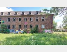 Zweistöckiges, verlassenes Backsteingebäude mit beschädigten Fenstern und Graffiti an den Wänden, umgeben von überwuchertem Gras und Bäumen. Im Hintergrund sind weitere einstöckige Gebäude und blauer Himmel sichtbar.
