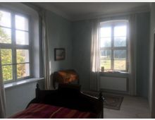 Helles Schlafzimmer mit zwei großen Fenstern, durch die Tageslicht einfällt. Im Raum befinden sich ein Bett, ein kleiner Schreibtisch aus Holz und ein Teppich auf Holzboden; vor den Fenstern hängen weiße Vorhänge.