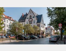 Straßenszene in einer Stadt mit einem historischen Gebäude im Stil der frühen Moderne, gekennzeichnet durch ein steiles schwarzes Dach und verzierte Fensterrahmen. Im Vordergrund stehen geparkte Autos entlang einer Pflasterstraße, umgeben von Bäumen und Verkehrsschildern.
