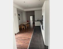 Langer, heller Innenraum mit einer offenen Küche und Essbereich. Links befindet sich ein Holztisch mit kleinen Stühlen auf einem Parkettboden, rechts ist eine moderne Küchenzeile auf einem dunkel gefliesten Boden zu sehen.