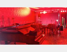 Ein Veranstaltungsraum mit roter Beleuchtung, ausgestattet mit einer kleinen Bühne im Hintergrund, Mikrofonständern und einer Seitenwand mit gemustertem Tapetendekor. Im Vordergrund steht ein Klavier, umgeben von Stehtischen mit Hockern auf einem Holzfußboden.