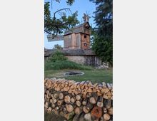 Alte hölzerne Windmühle mit Flügeln und Schindeldach hinter einem niedrigen Nebengebäude auf einem ländlichen Grundstück. Im Vordergrund liegen gestapelte Brennholzscheite, daneben eine steinerne Feuerstelle, umgeben von Wiese und Bäumen.