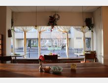 Innenraum eines Cafés mit rustikalem Holztisch im Vordergrund und mehreren Stühlen. Durch große Fenster mit transparenten Vorhängen sieht man einen Außenbereich mit einem Wohnhaus, Blumenbeeten und einer bunten Neon-Dekoration in Form einer Katze.