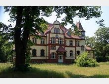 Historisches Gebäude mit gelber Fassade und roten Akzenten im Stil der Gründerzeit, umgeben von Bäumen und einer Wiese. Das Gebäude hat verzierte Fenster, ein spitzes Dach und abgerundete Giebel.