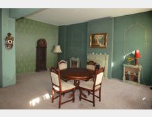 Ein klassisch eingerichtetes Wohnzimmer mit grünen Wänden, einer dekorativen Tapete, einem runden Holztisch und vier gepolsterten Holzstühlen. Im Hintergrund befinden sich ein antiker Standspiegel, ein Kamin mit goldenen Akzenten, ein Gemälde sowie ein Regal mit Büchern und Teetassen.