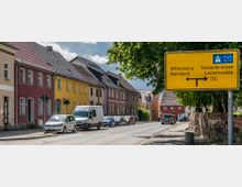 Straßenszene in einer Kleinstadt mit farbigen Reihenhäusern auf der linken Seite, parkenden Autos und einem großen gelben Wegweiser auf der rechten Seite, der auf Autobahn- und andere Ziele hinweist. Im Hintergrund sind Bäume und weitere Gebäude sichtbar.