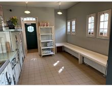 Kleiner Raum mit beigem Fliesenboden, einer weißen Wandbank entlang eines Heizkörpers und mehreren kleinen Fenstern mit Blumenkästen. Links steht eine Glasvitrine und ein Regal mit Produkten, hinten führt eine grüne Tür nach draußen.