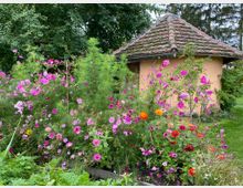 Kleiner Garten mit bunten Blumen, darunter pinke, rote und orangefarbene Blüten, vor einem runden Gebäude mit beigem Putz und einem Dach aus alten Ziegeln. Im Hintergrund sind Bäume und dichtes Grün sichtbar.