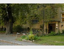 Mehrfamilienhaus mit verwitterter Fassade, umgeben von Bäumen und einem kleinen bepflanzten Vorgarten mit Holzzaun. Fahrräder stehen vor dem Gebäude, dessen Eingangstür leicht geöffnet ist.