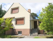 Einfamilienhaus mit gelber Fassade und braunem Sockel, Satteldach, geschlossenen Rollläden und gemauerten Fensterrahmen. Eine Treppe führt zum überdachten Eingangsbereich, umgeben von Garten mit Bäumen und Grünflächen.